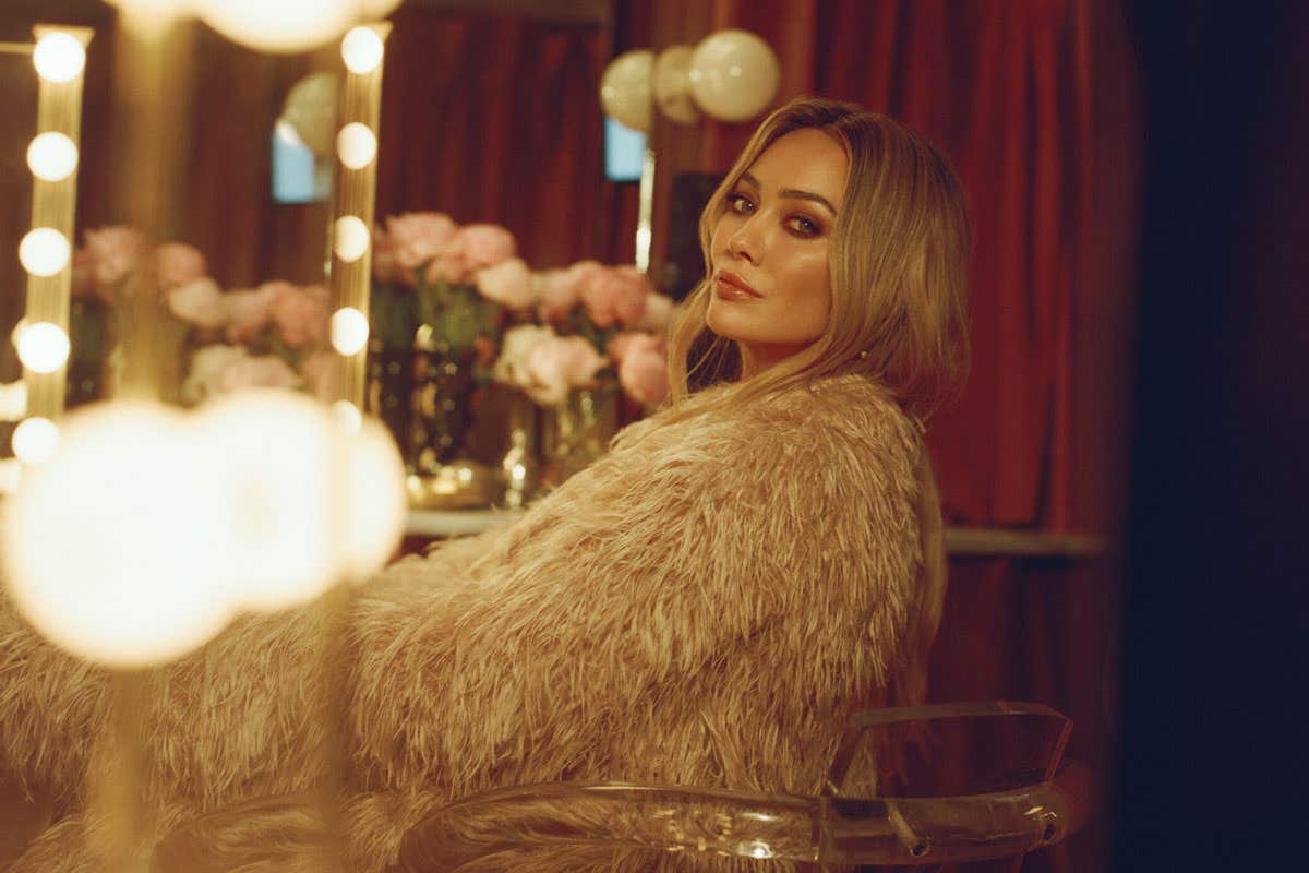 Hilary Duff sitting on clear seat in a fur coat in front of a vanity table looking towards the camera. 