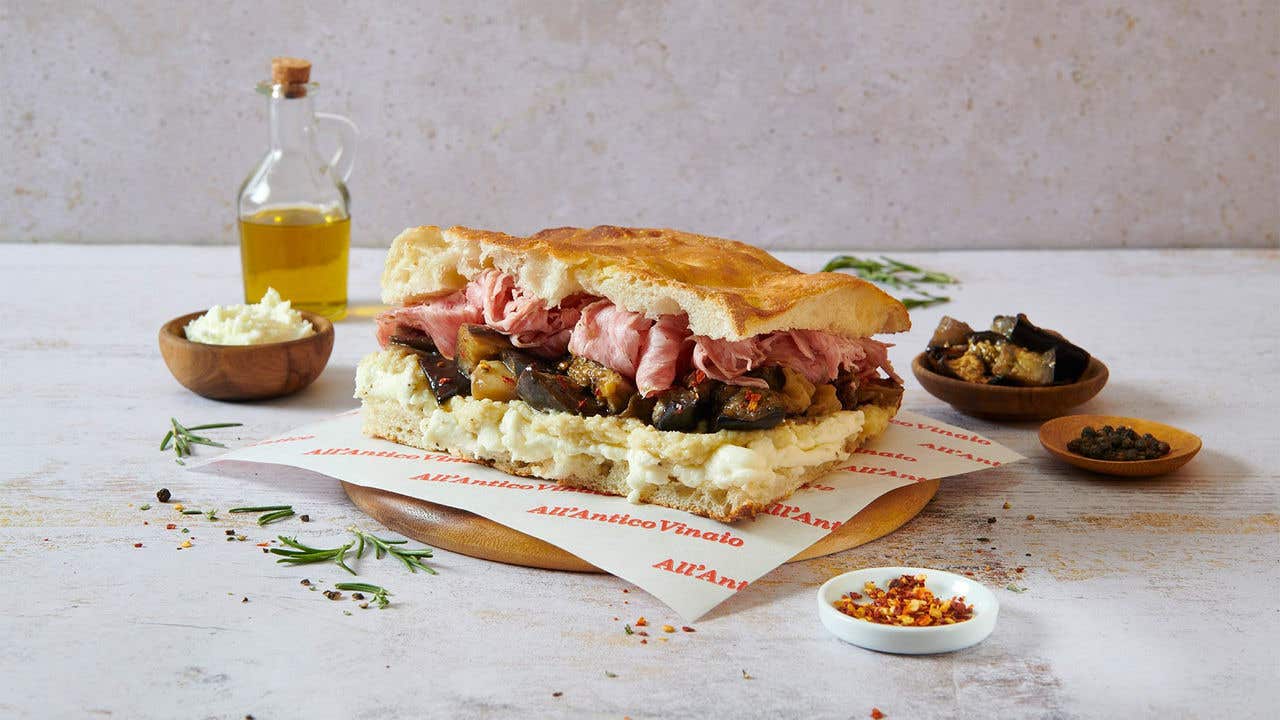 Panini sandwich with ham and olive tapenade on a round wood plate surrounded by seasonings and a bottle of olive oil. 