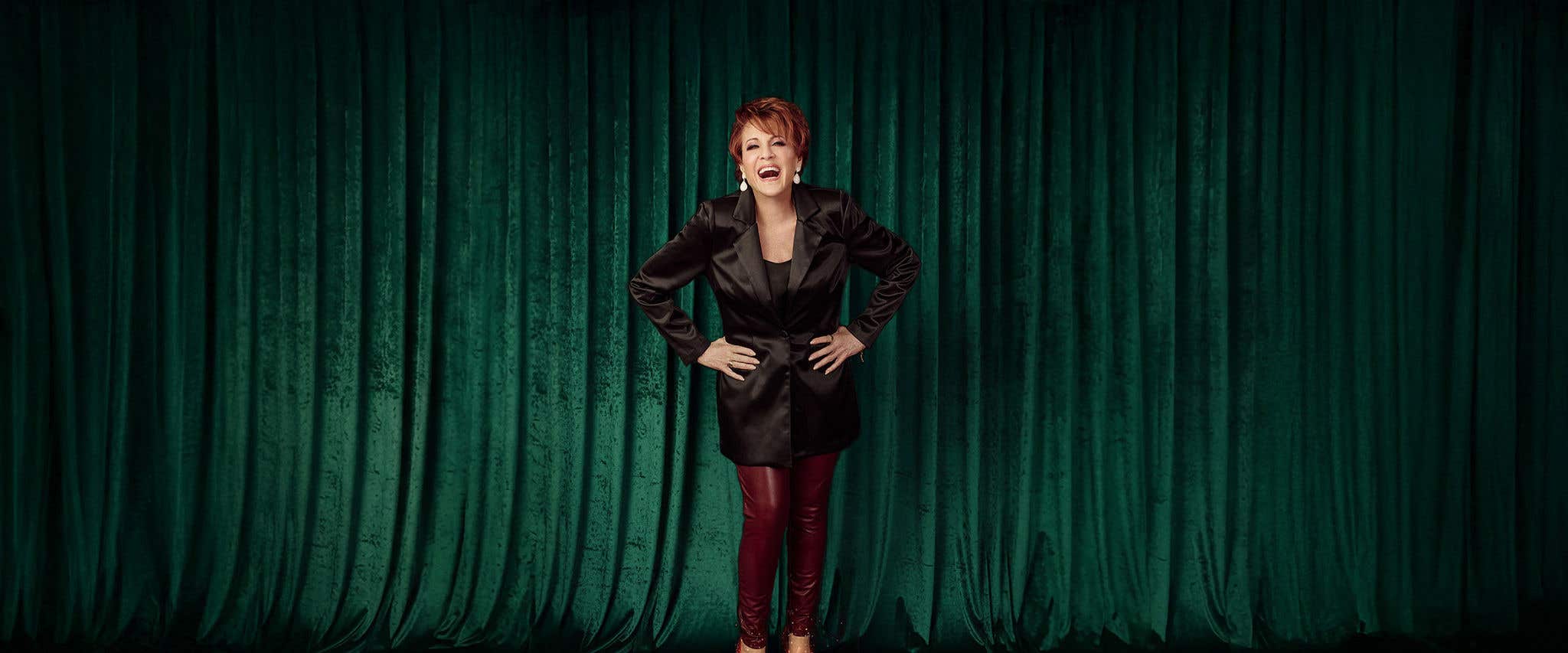 Lorna Luft standing in front of green curtains laughing wearing a black top with black jacket, burgundy pants, and ruby red shoes. 
