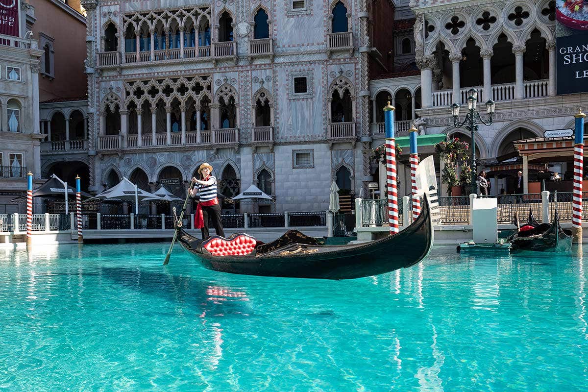 A classic Venetian gondola with red leather seats floats on vibrant teal water. The gondola is piloted by a gondolier dressed in a blue and white striped shirt, red sash tied into a bow, red handkerchief, black pants, and tan brimmed hat.