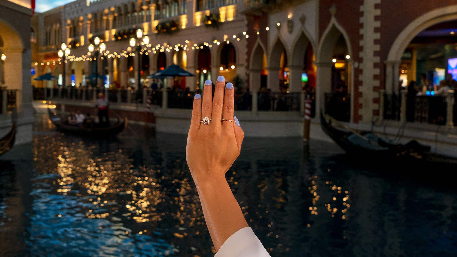A woman’s left hand with an engagement ring on her ring finger and purple painted nails in front of the Grand Canal in the Grand Canal Shoppes.