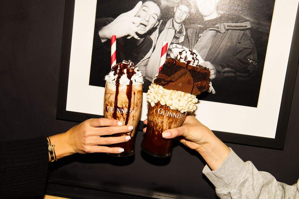 Two hands holding milkshakes topped with whipped cream, chocolate drizzle, and brownies in front of a black and white photo.