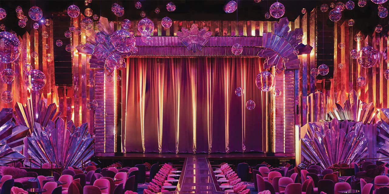 Vibrant theater with pink seating, a long runway, shimmering crystal décor, hanging orbs, and a glowing curtain-lit stage.