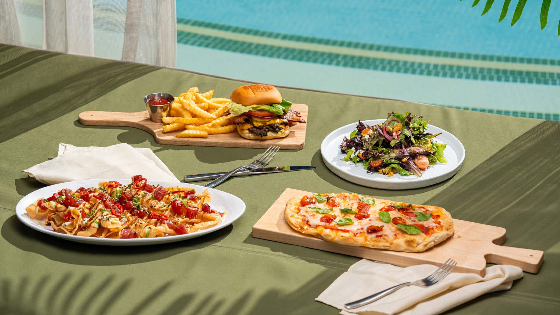 Poolside lunch spread with a burger and fries, mixed salad, flatbread pizza, and nachos.
