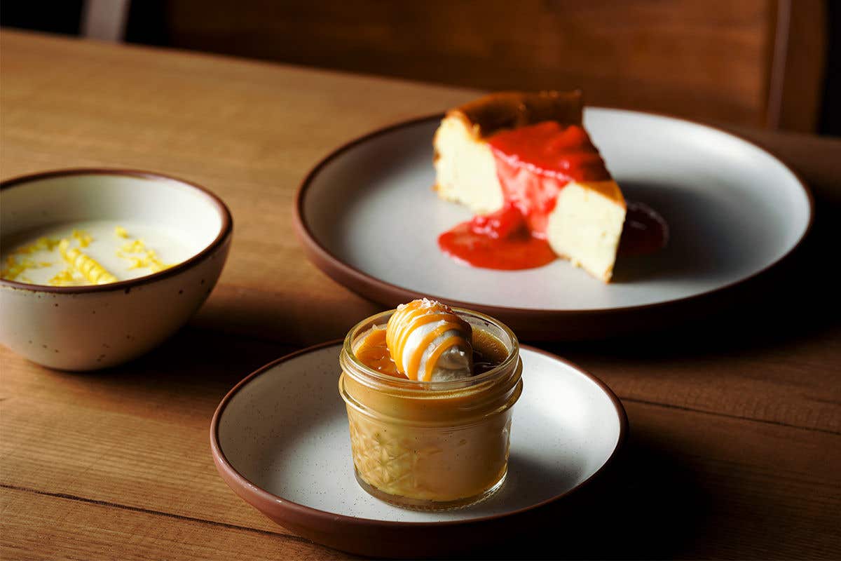 Three desserts on table, a butterscotch pot de crème topped with caramel and ice cream, a slice of cheesecake topped with a red sauce and bowl of panna cotta topped with lemon.