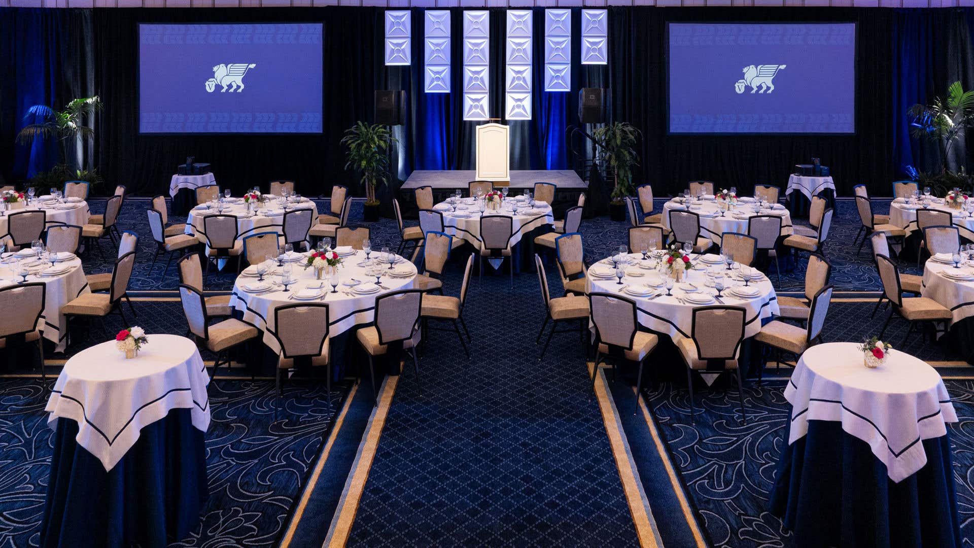Elegant ballroom with round tables in navy and white linens, blue accent lighting, two projection screens, and a staged podium.