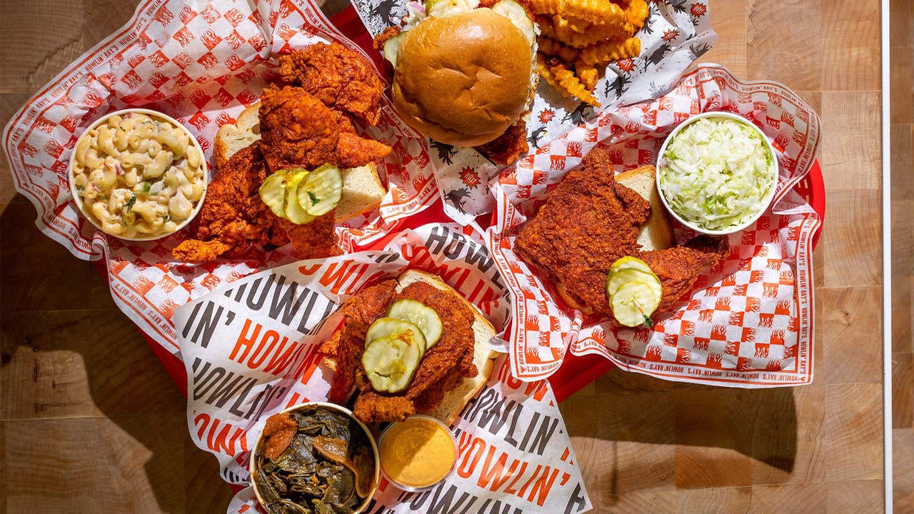 Four baskets with paper liners in them grouped together containing fried chicken with pickles on top and sides of collard greens, cheese and cole slaw.