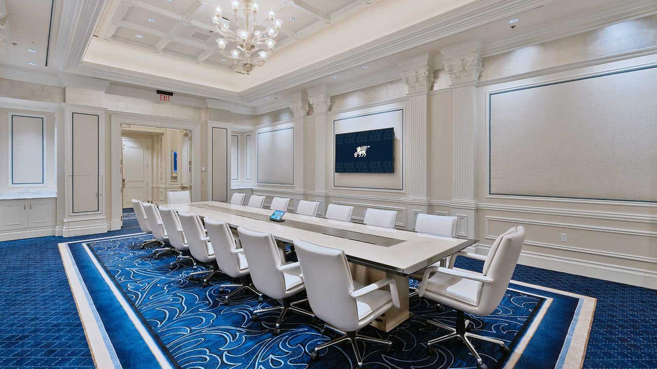 A table surrounded by chairs staged for a meeting set-up in a boardroom with a chandelier, blue carpet and three TVs hung on white walls.