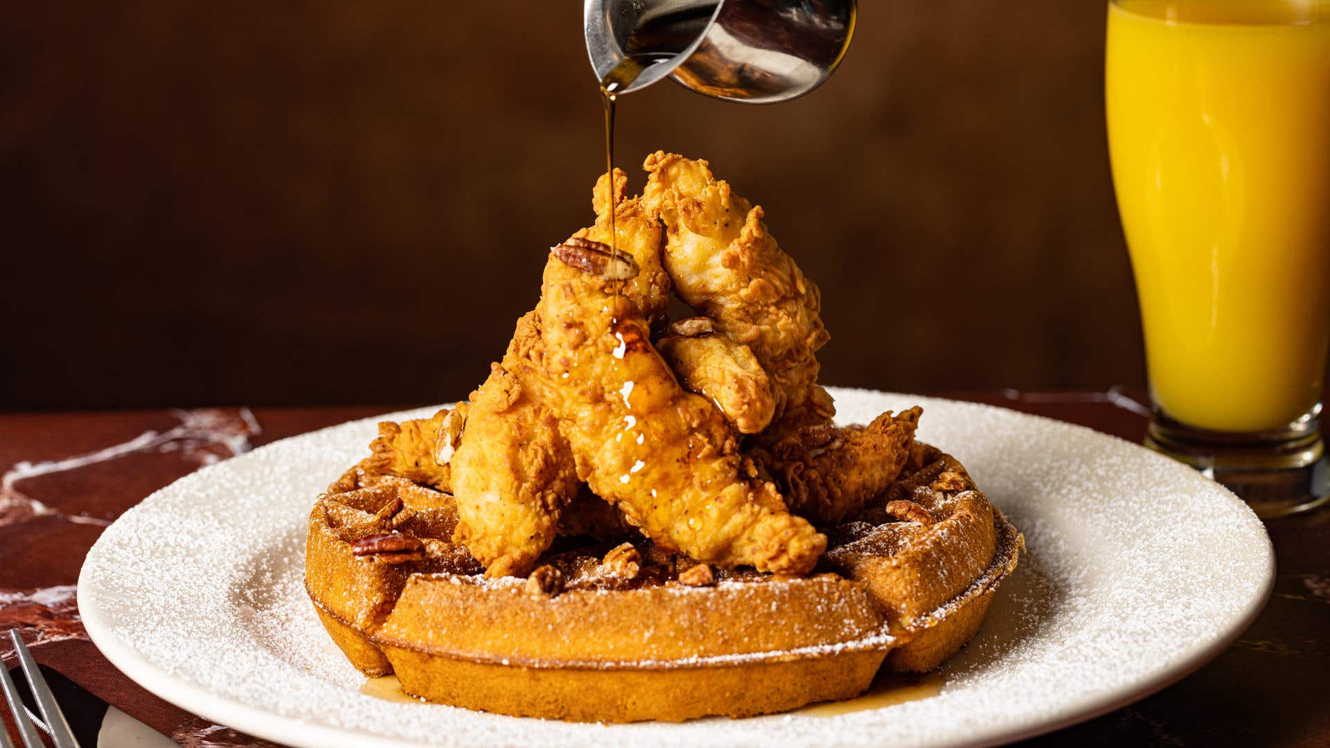 A white round plate with Belgian waffle topped with breaded chicken as syrup is poured over the top.